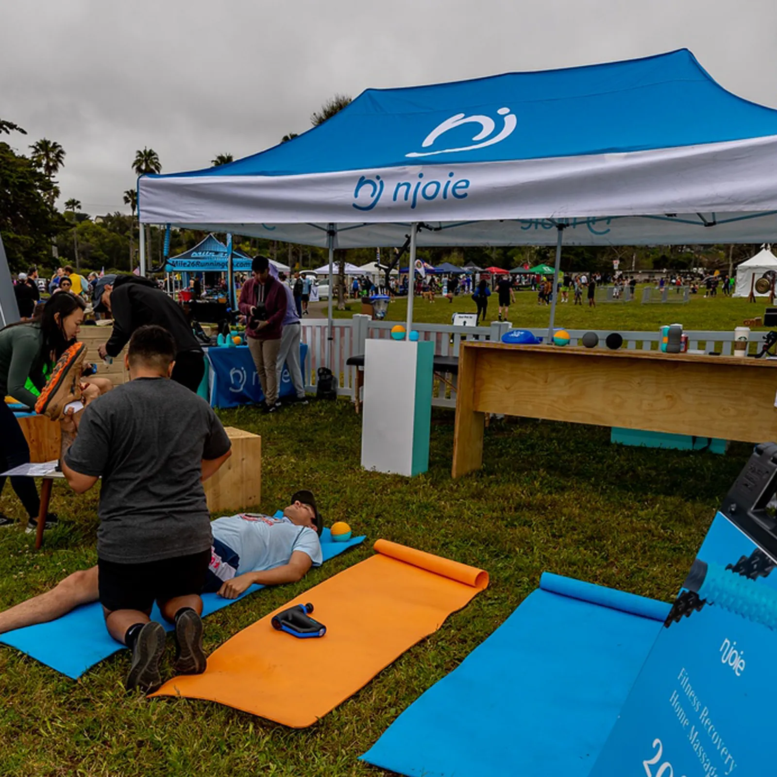 People receiving massage or recovery treatment on mats at a race event, with a sponsor tent in the background.