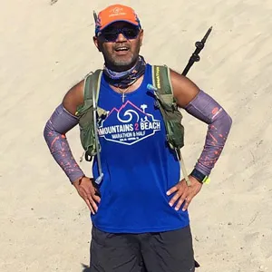 A male runner smiles at the camera, wearing a blue Mountains 2 Beach Marathon & Half t-shirt and a hydration pack.