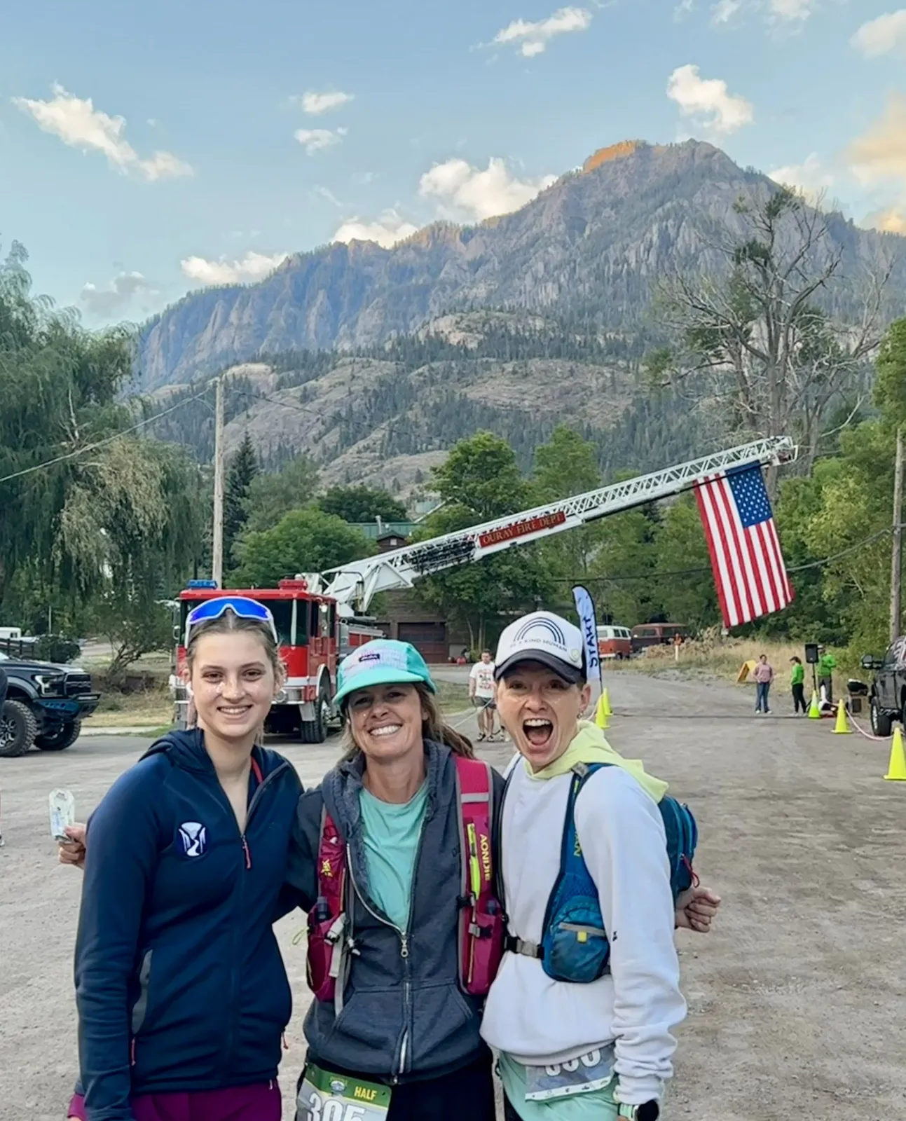 Three smiling women pose for a photo before the Mt. Sneffels Half Marathon, with a fire truck and American flag behind them, and a scenic mountain range in the background.