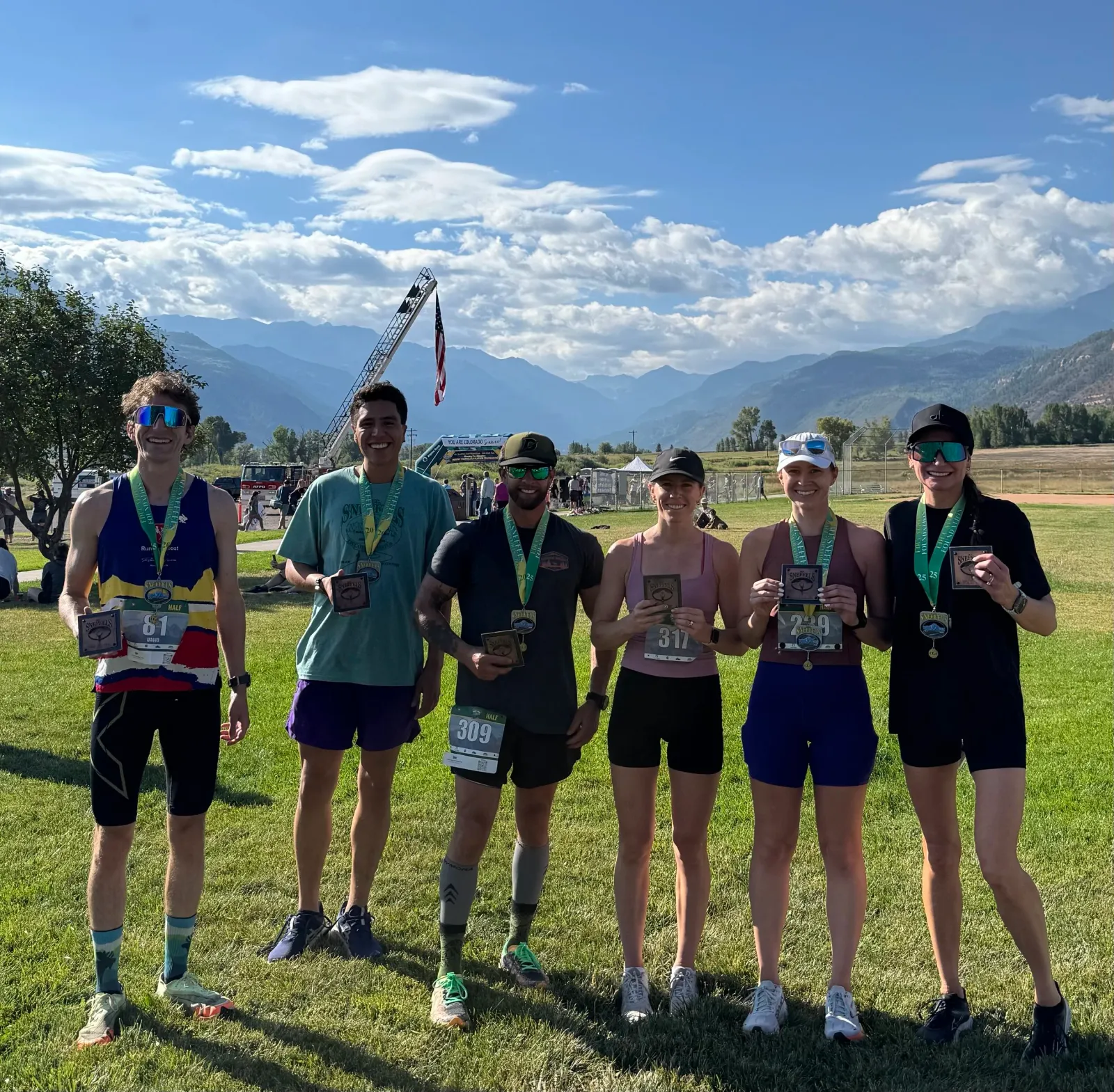 A group of six runners, winners of the Mt. Sneffels Half Marathon, pose for a photo with their medals and awards, with a scenic mountain range in the background.