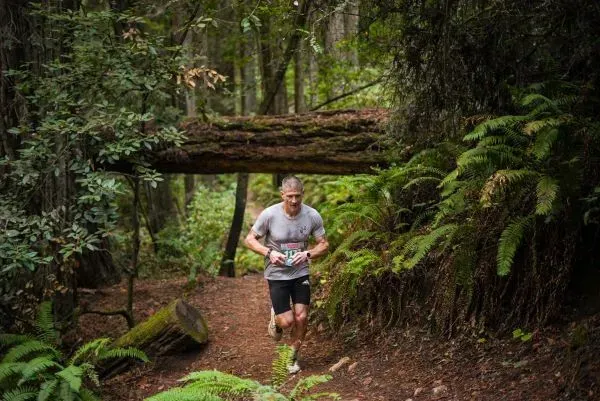 A male runner runs on a dirt trail through a lush, green forest, passing under a fallen log.