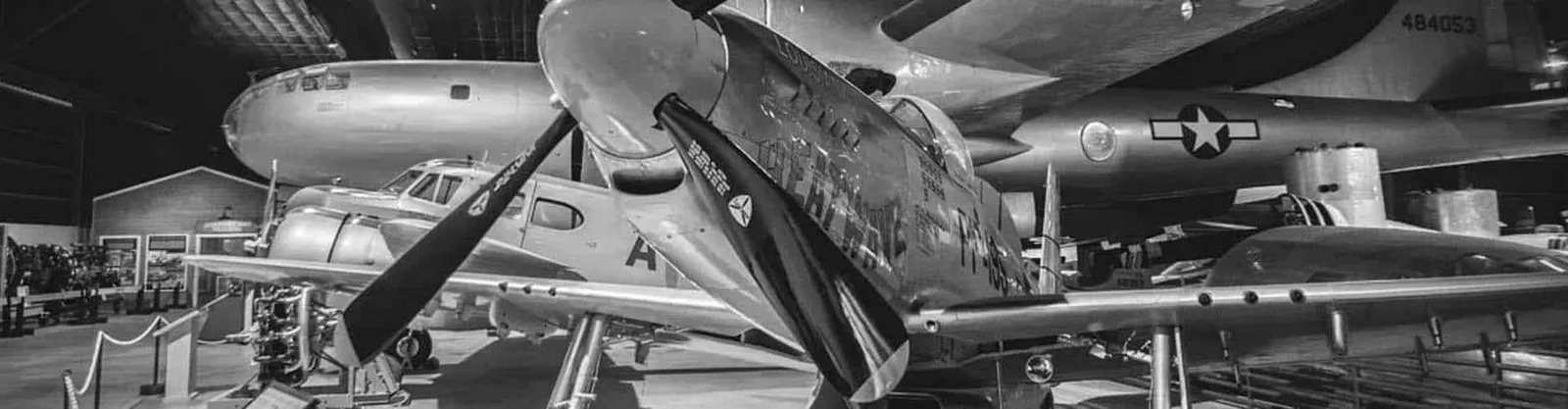 A black and white photograph of several vintage airplanes, including a prominent fighter plane in the foreground and a large bomber in the background, displayed indoors.
