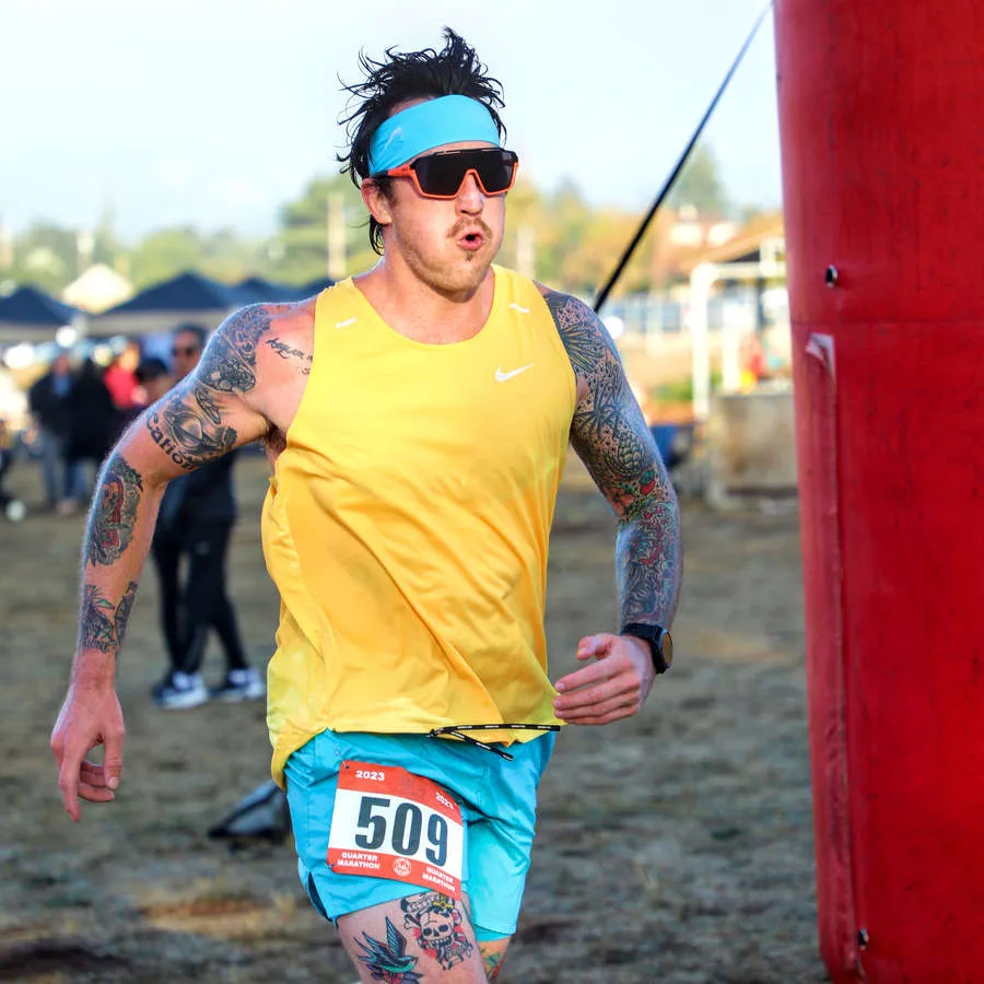 A male runner with tattoos, wearing a yellow tank top and blue shorts, runs past a red archway during the Napa Half Marathon.