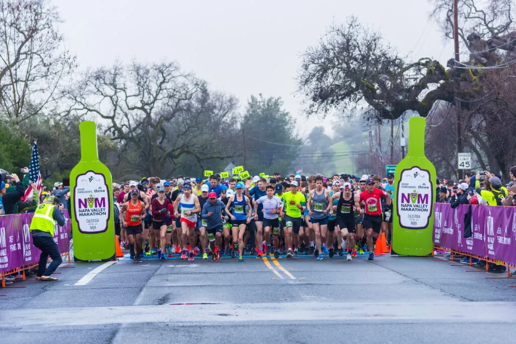 Runners at the start line of the Napa Valley Marathon, with large green wine bottle cutouts on either side.