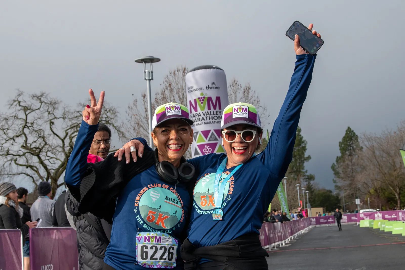 Two smiling women in race attire, one holding up a phone and the other making a peace sign, celebrate at the Napa Valley Marathon.