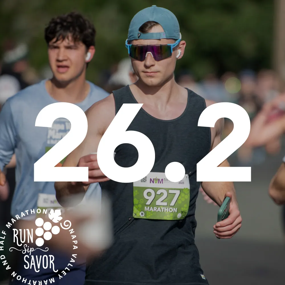 Two male runners on a road race course, with a large white overlay of '26.2' and race branding.