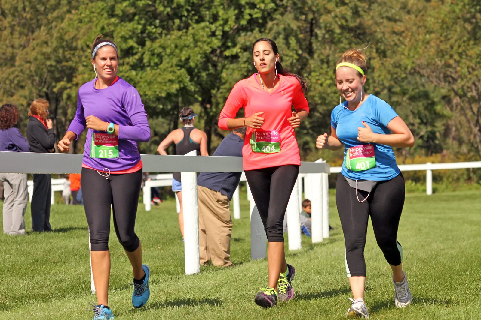 Three female runners in colorful shirts and black leggings running on a grassy course, with trees in the background.