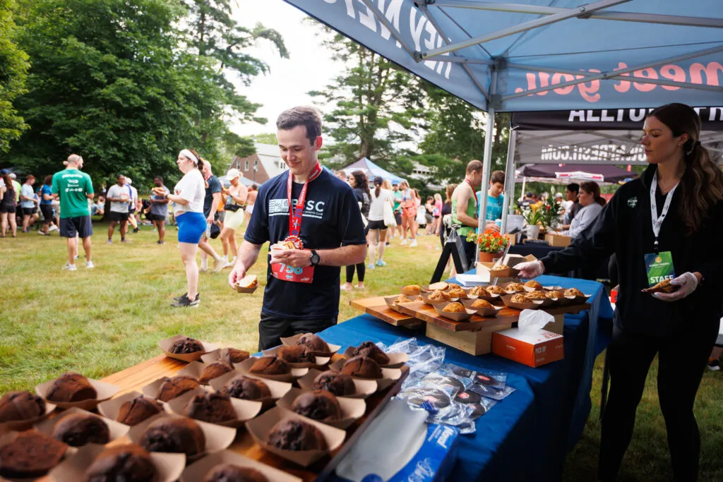 A man in a race bib takes a muffin from a table, with other runners and volunteers in the background at an outdoor event.