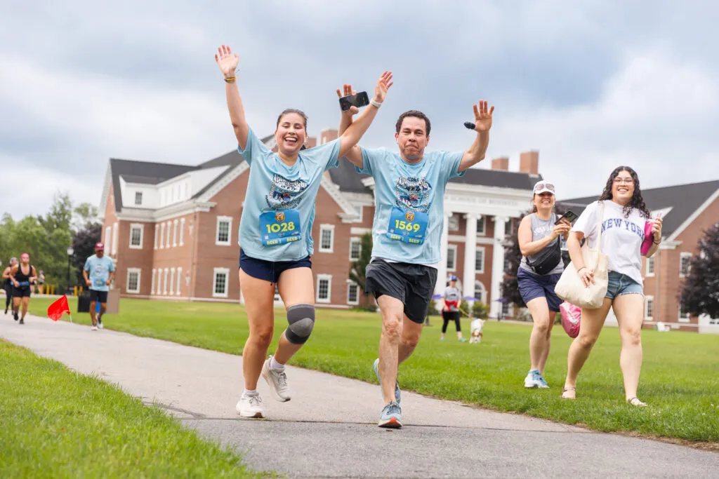 Two runners with their arms raised in celebration as they run towards the camera, with other participants and a large building in the background.