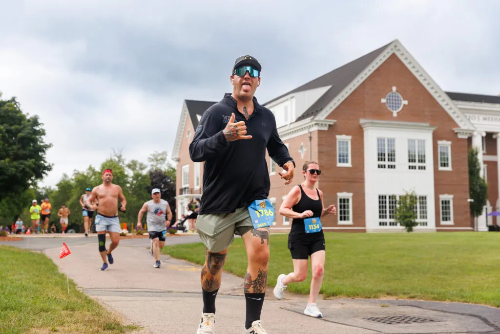 A runner with tattoos and sunglasses sticks his tongue out while running, with other runners and a brick building in the background.