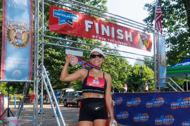 A smiling female runner wearing sunglasses and a white hat holds up her race medal at the finish line, with a 'FINISH' banner overhead.