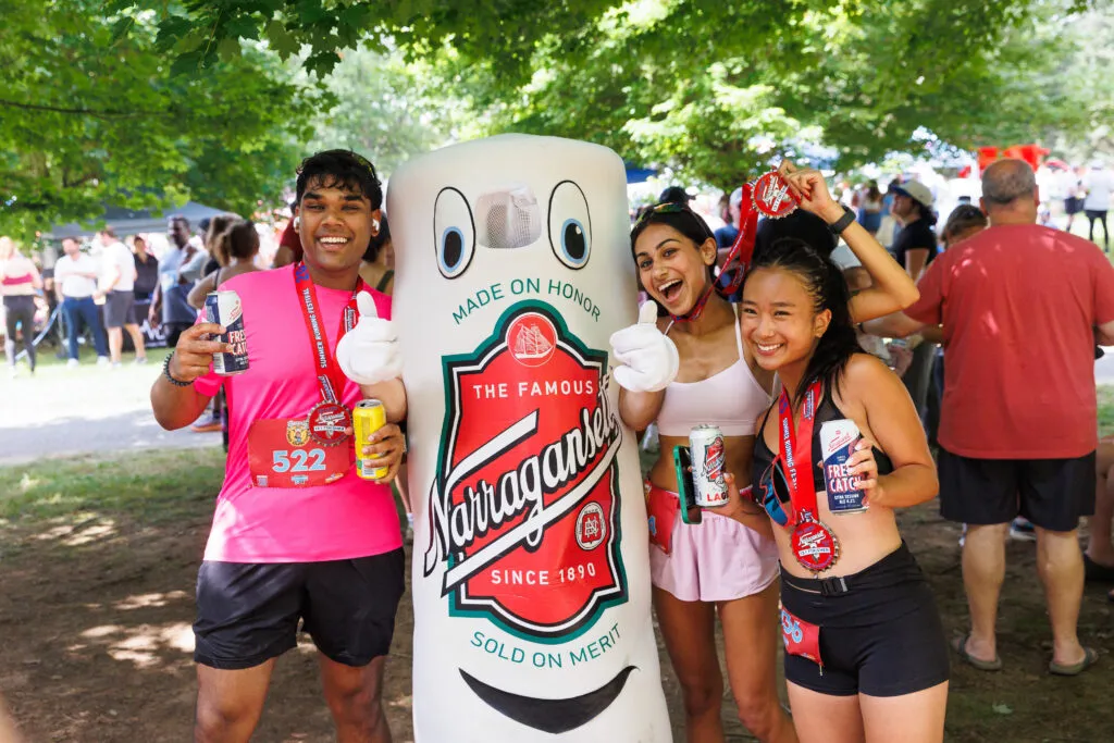 Three smiling runners pose with a Narragansett beer mascot, holding medals and drinks after completing a race.