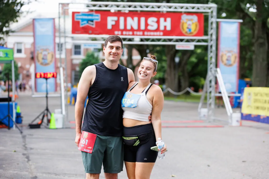 A smiling couple poses for a photo at the finish line of a road race, with the finish banner visible behind them.