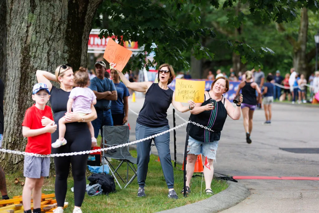 Spectators cheer for runners at the Narragansett Summer Running Festival, holding signs that say 'Falvey!' and 'You're Amazing Falvey!'
