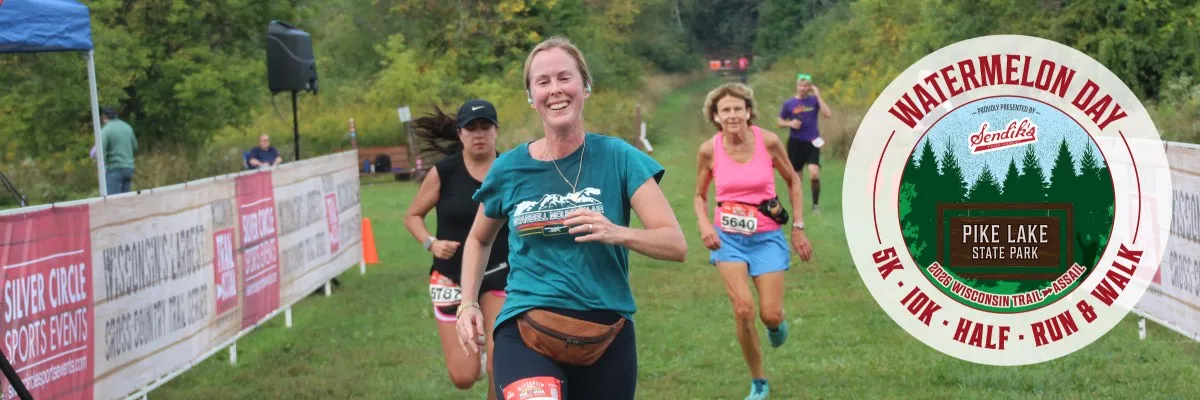 A smiling female runner leads a group of participants on a grassy course, with a race logo visible on the right.
