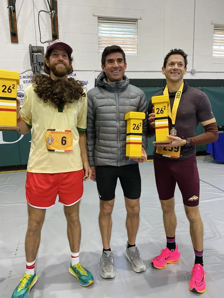 Three male runners smiling and holding awards, one wearing a medal, after a race.