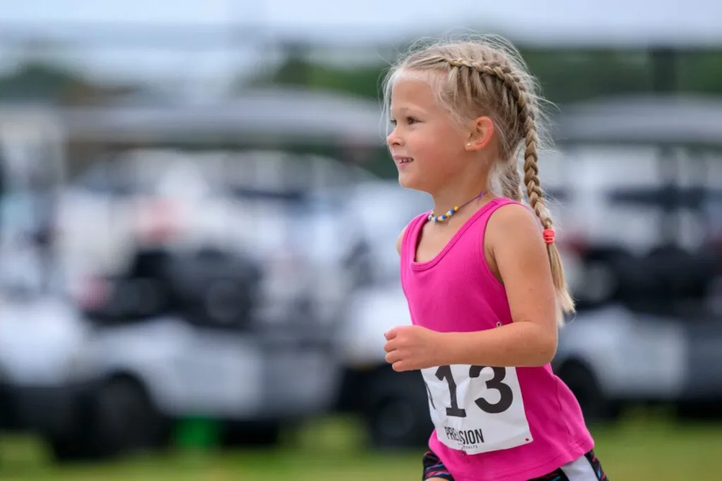 A young girl with braided blonde hair wearing a pink tank top and bib number 13 runs in a race.