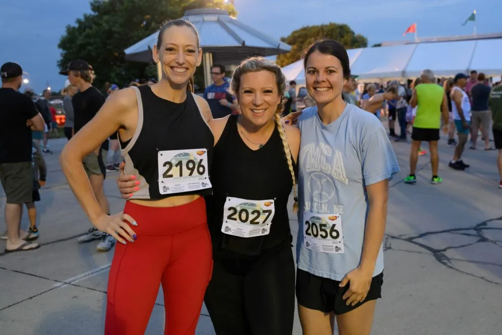 Three smiling women wearing race bibs pose for a photo at the start of a race, with tents and other runners in the background.