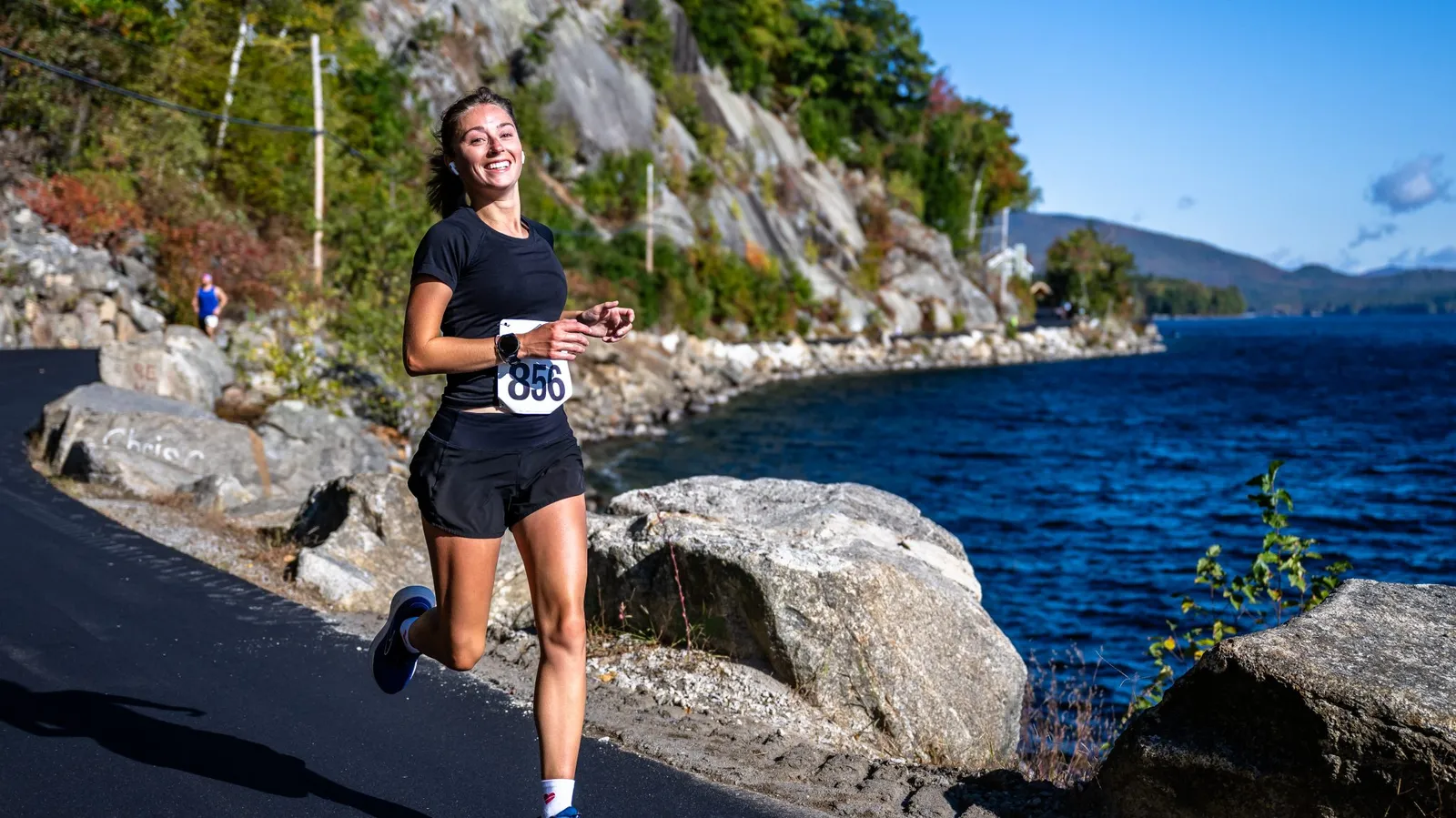 A female runner smiles as she runs along a road next to a large blue body of water, with rocky hills and trees in the background.