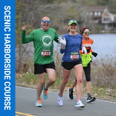 Two runners smile and give a peace sign while running along a road next to a body of water, with a third runner behind them.