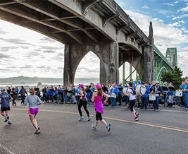 Runners pass under a large bridge while a band plays music on the side of the road.
