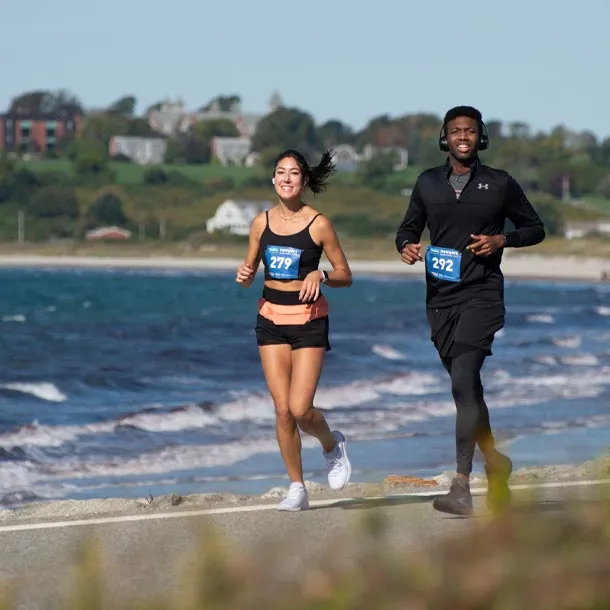 A male and female runner are running along a paved path next to the ocean. The female runner is in the foreground on the left, wearing a black tank top and shorts, with bib number 279. The male runner is to her right, wearing a black long-sleeved shirt and shorts, with bib number 292. Both runners are smiling and appear to be enjoying the race. The ocean is to their left, and buildings are visible on a hill in the background.