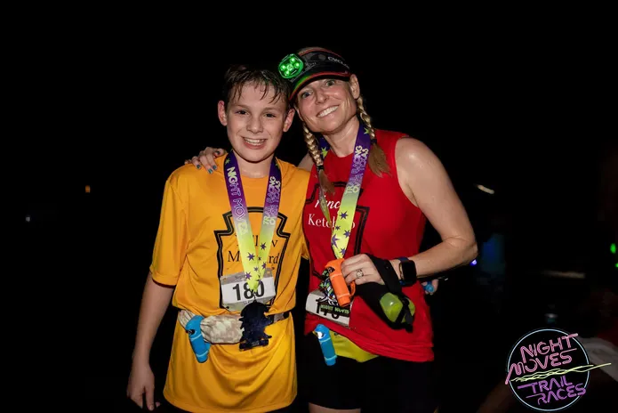 A young boy and an adult woman smile at the camera, both wearing race medals and bibs, after completing a night trail race.