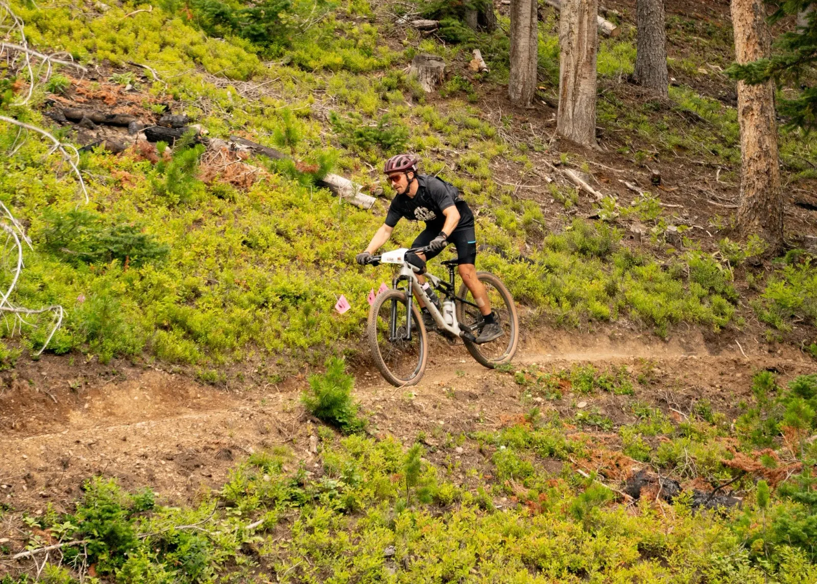 A male mountain biker wearing a helmet and sunglasses rides on a dirt trail through a forest.