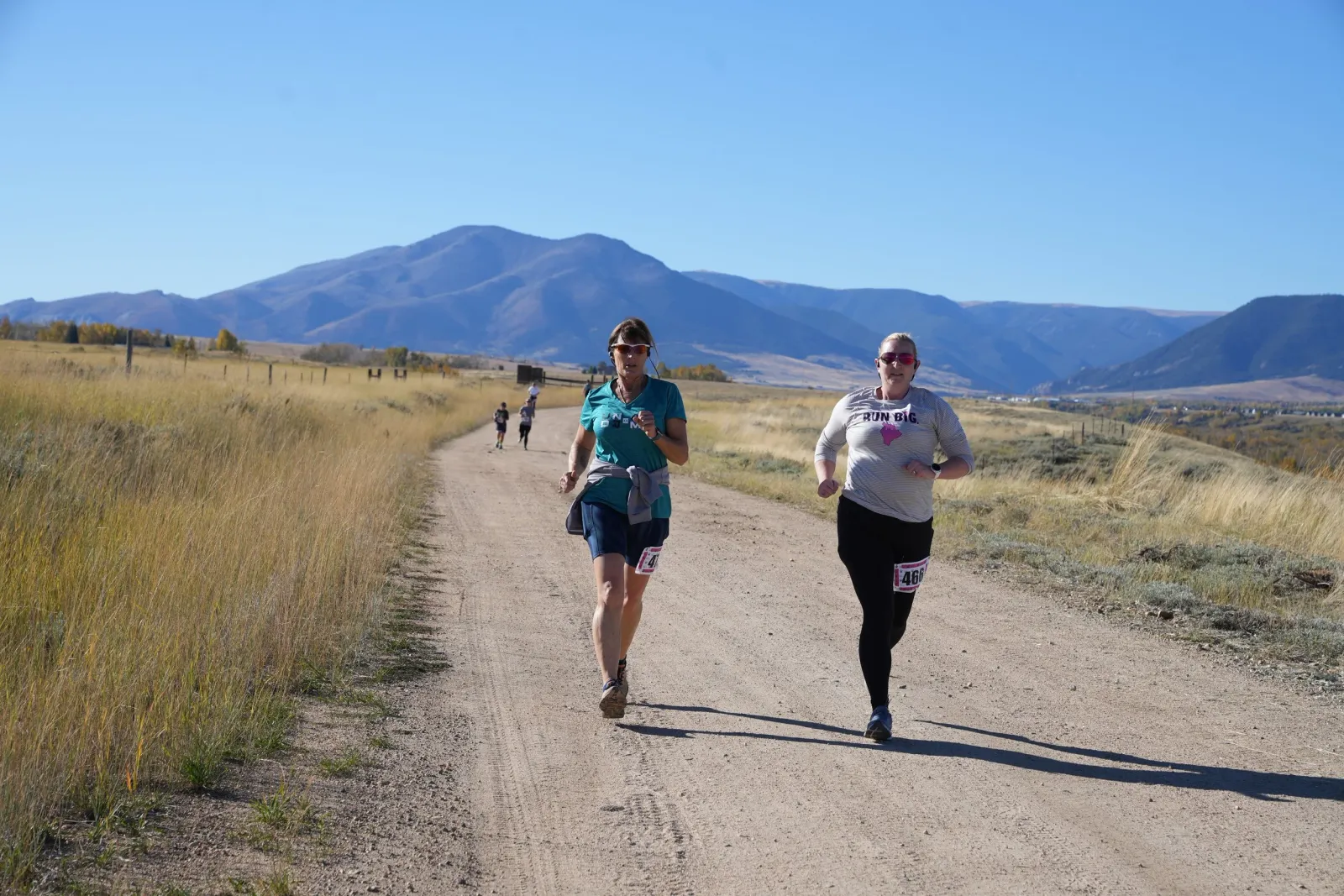 Two women running on a dirt road with mountains in the background.