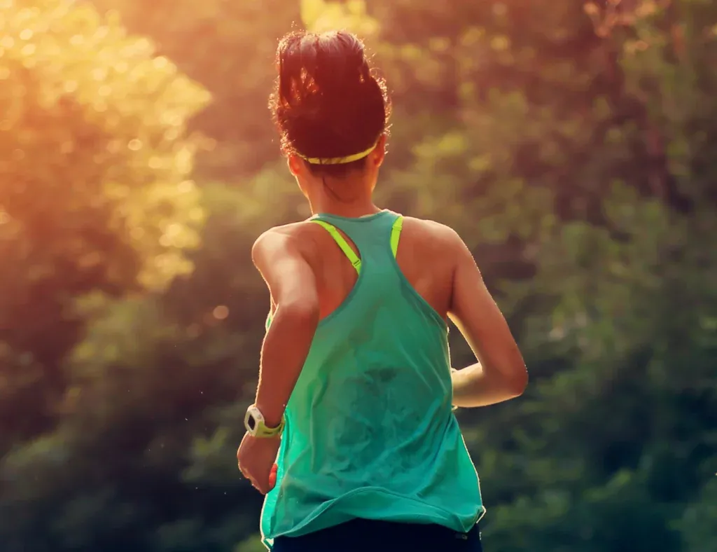 A female runner seen from behind, running on a road with trees and sunlight in the background.