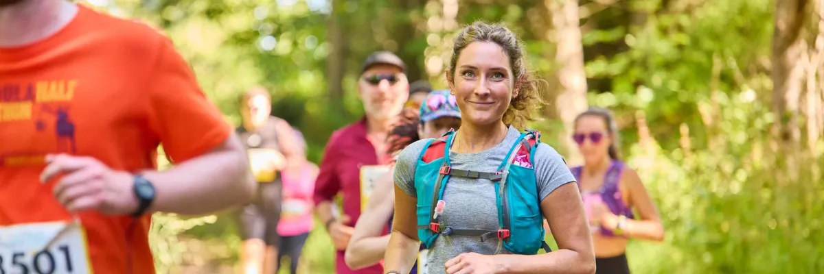 A woman smiles at the camera while running on a wooded trail, with other runners behind her.
