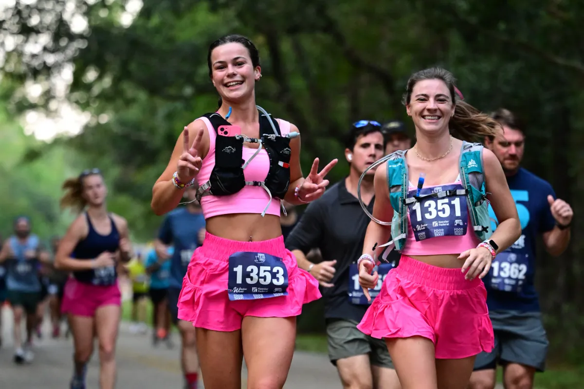 Two female runners in pink outfits smiling and giving peace signs while running a race.