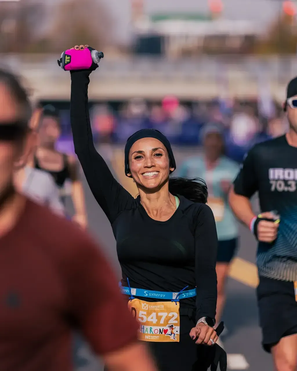 A female runner with her arm raised in celebration, holding a pink water bottle, smiles broadly during a road race.