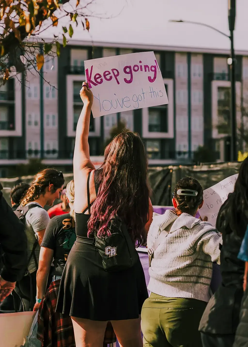 A person holds up a sign that says 'Keep going! You've got this!' at a race event, with other spectators in the background.