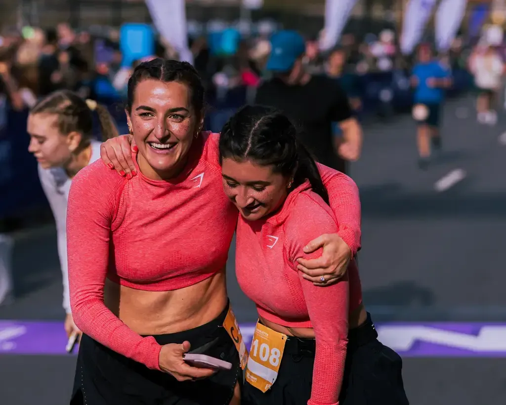 Two women in pink running tops embrace and laugh, with race bibs visible, at what appears to be the finish line of a road race.