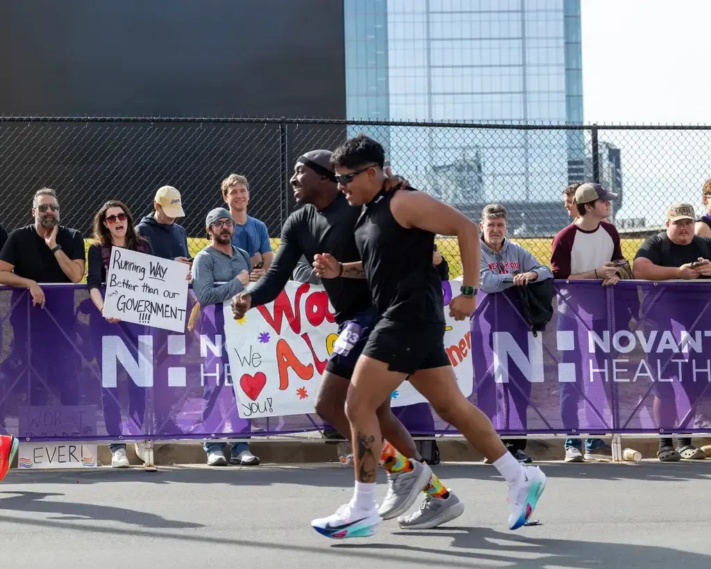 Two runners are shown in profile, with one runner's arm around the other's shoulder, as they run past spectators holding signs.