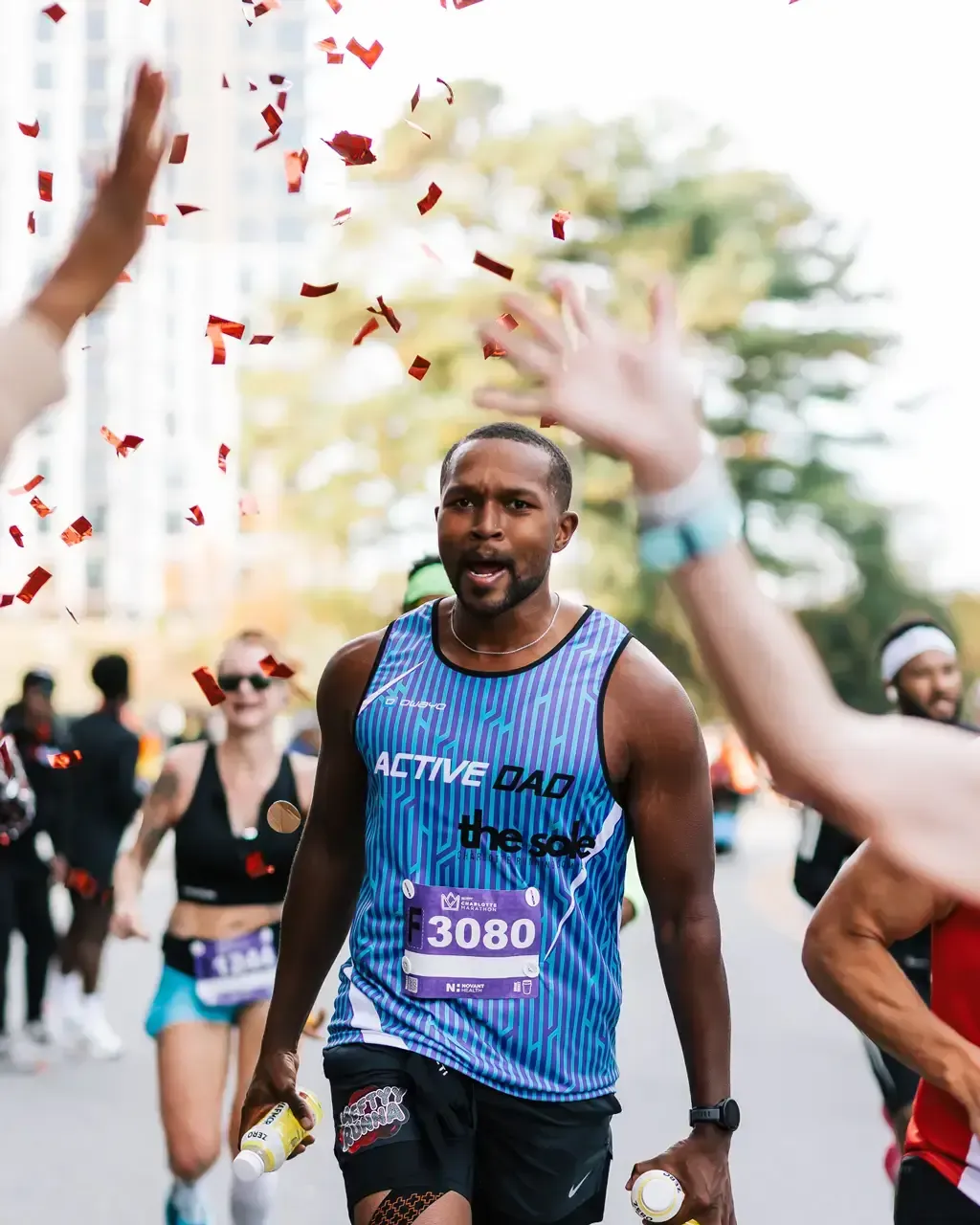 A male runner with a bib number F3080 crosses the finish line with confetti falling around him and hands reaching out to high-five him.