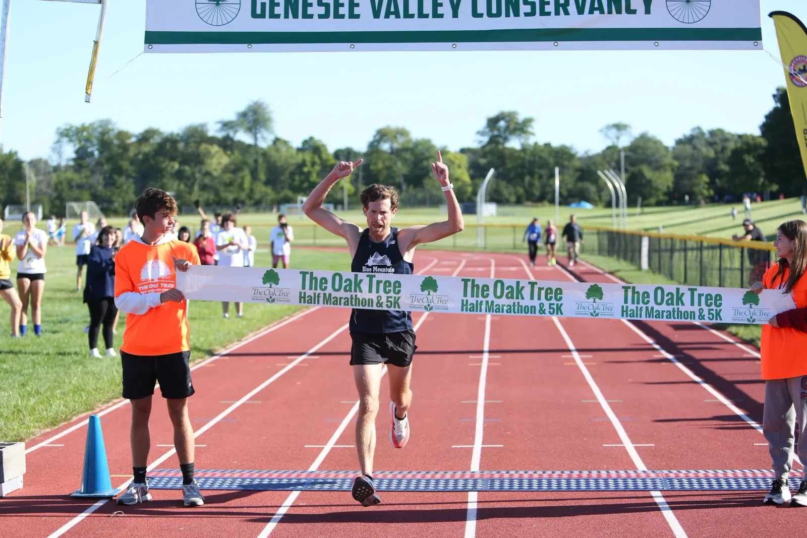 A male runner crosses the finish line with his arms raised in celebration, with a banner that reads "The Oak Tree Half Marathon & 5K" overhead.