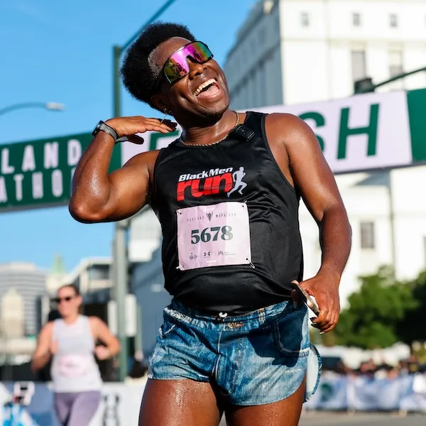 A joyful runner in a black 'Black Men Run' tank top and denim shorts crosses the finish line, with sunglasses reflecting the sky.