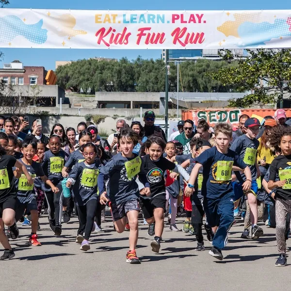 Children running at the start of a Kids Fun Run, with a banner overhead that says 'EAT. LEARN. PLAY. Kids Fun Run.'