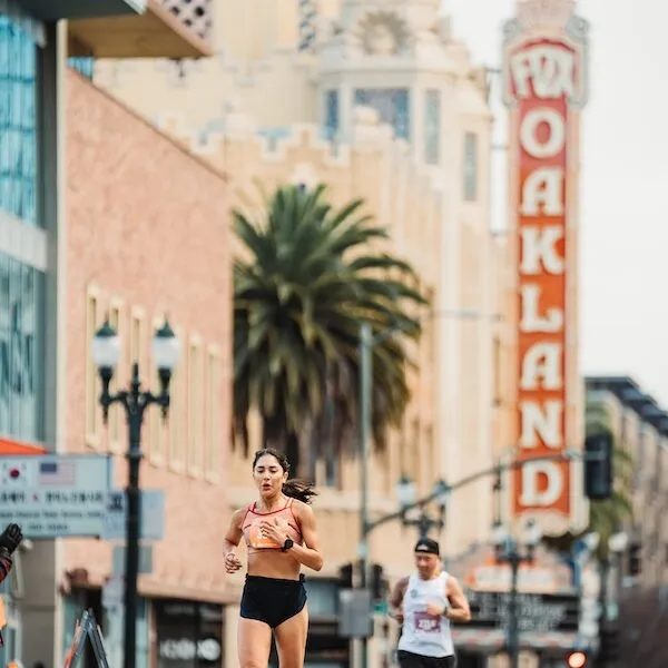 A female runner in a sports bra and shorts runs down an urban street with the historic Fox Theater in Oakland visible in the background.