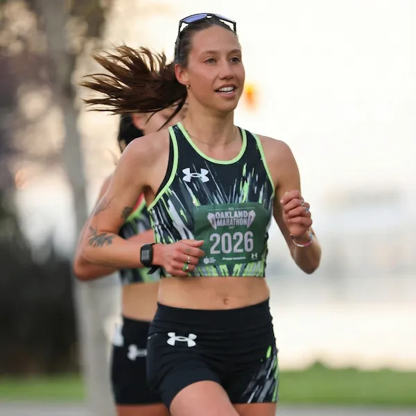 A female runner with her hair blowing in the wind, wearing a green and black patterned tank top and black shorts, running in a race.