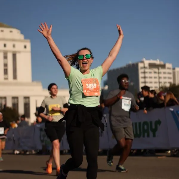 A female runner with arms raised in celebration, wearing sunglasses and a green t-shirt with bib number 3889, running on a road with other participants and spectators in the background.