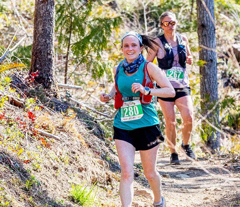 Two female runners on a wooded trail, one in the foreground wearing a teal shirt and bib number 1280, smiling at the camera.