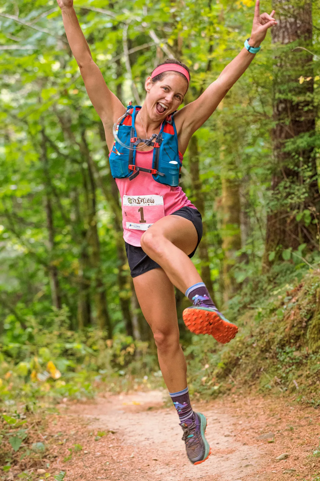 A female runner with her arms raised in celebration, wearing a pink shirt and bib number 1, jumping on a dirt trail surrounded by trees.