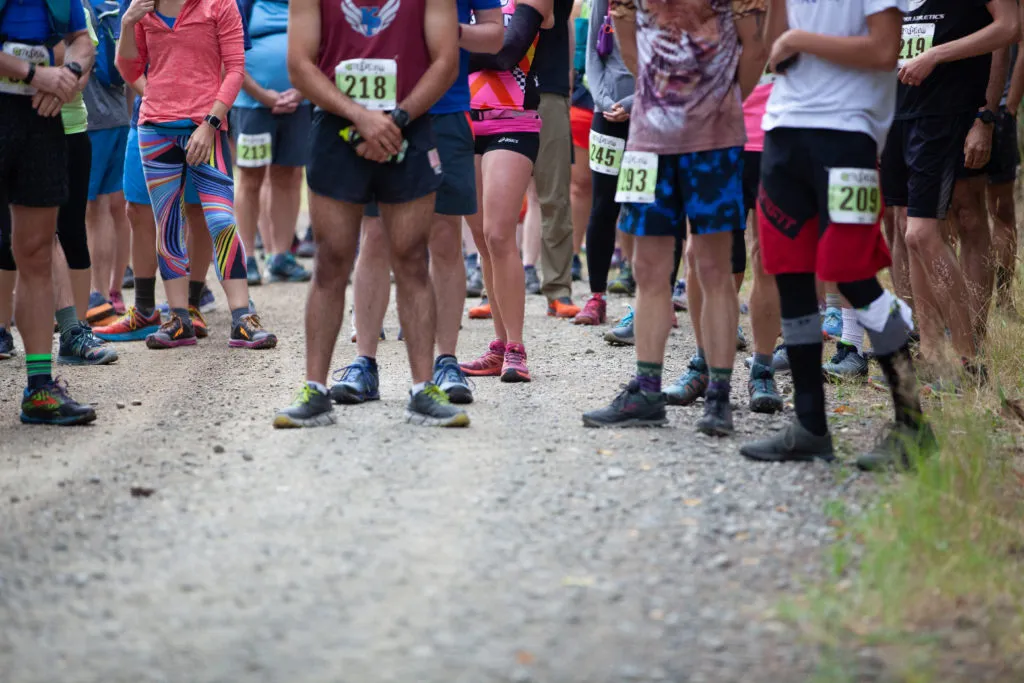 A group of runners stand at the start line, wearing bib numbers and athletic gear.