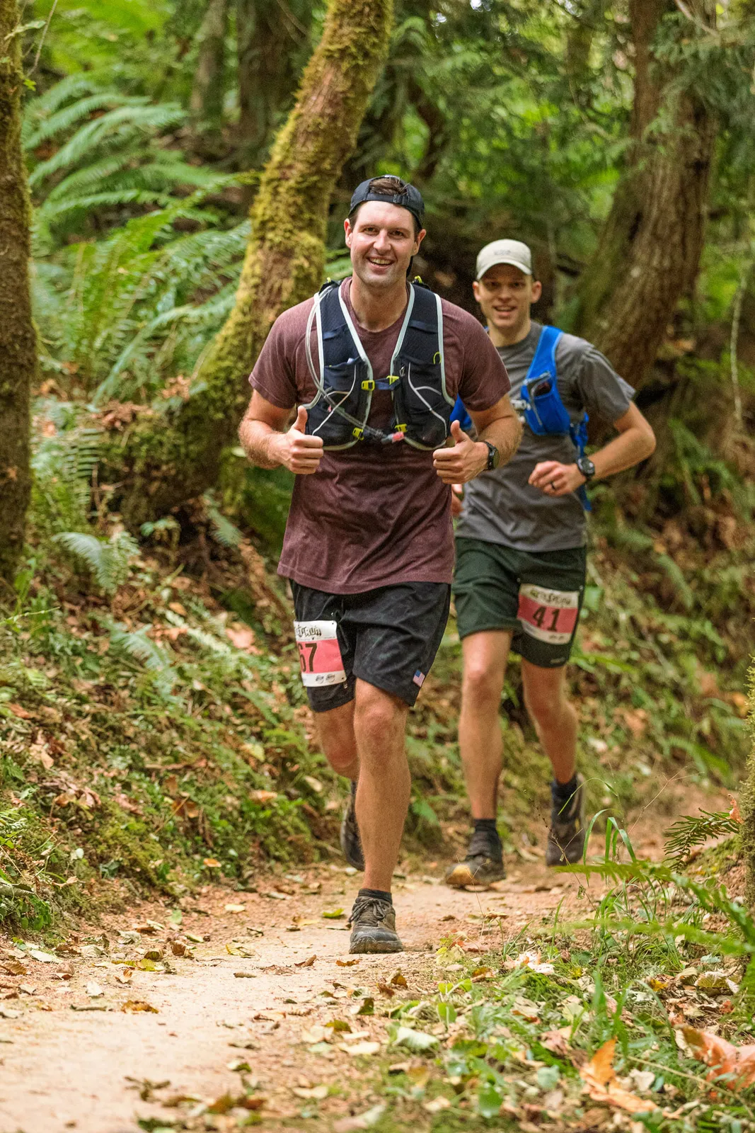 Two male runners smiling and giving thumbs up while running on a wooded trail during a race.