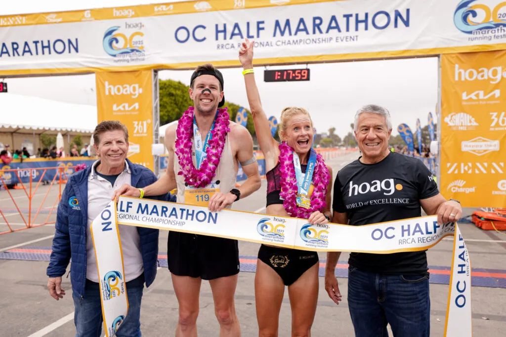Two runners, a man and a woman, celebrate crossing the finish line at the OC Half Marathon, holding a banner and wearing leis and medals.