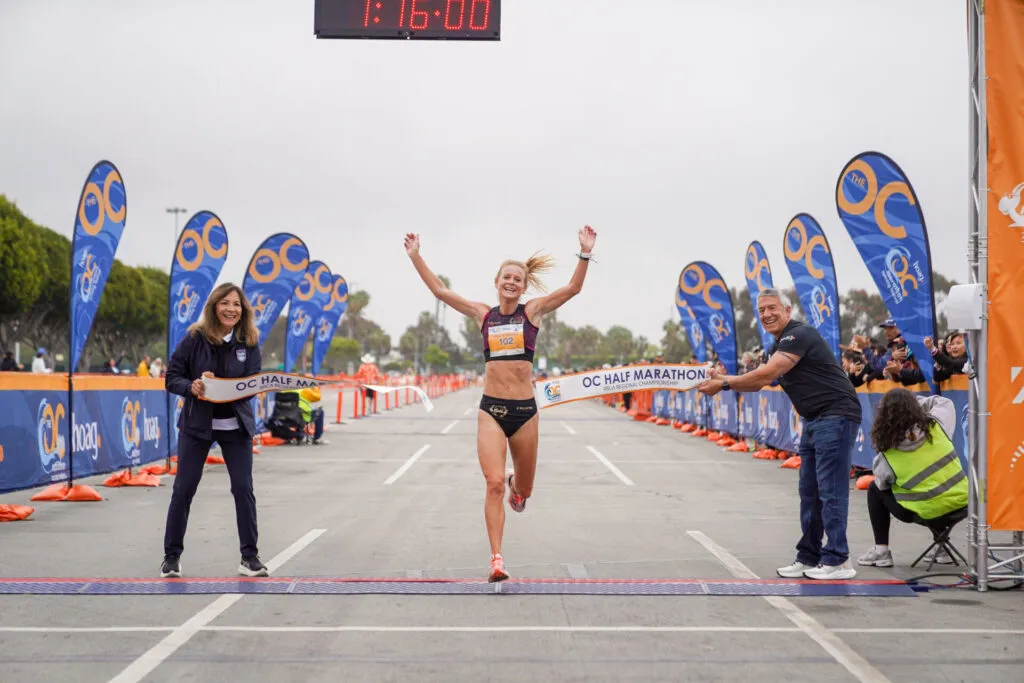 A female runner with her arms raised in celebration crosses the finish line of the OC Half Marathon, breaking a tape held by race officials, with spectators cheering in the background.