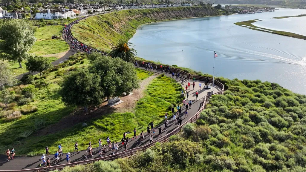 Runners on a paved path alongside a bay with green hills and houses in the background.